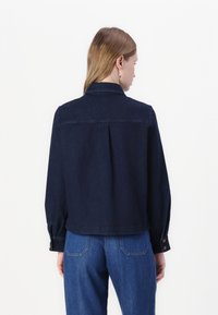 Giacca in denim scuro con colletto, polsini abbottonati e dettaglio plissettato sul retro, abbinata a jeans in denim blu.