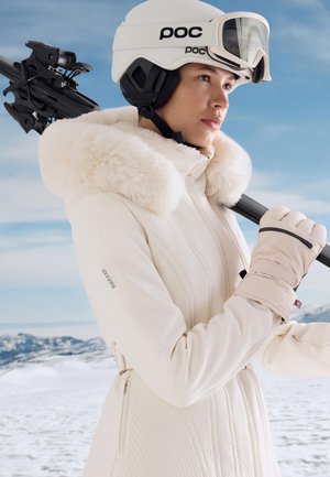 Hvid skijakke med pelsforet hætte, tekstureret design og lynlåse detaljer. Sort skihjelm, goggles og beige handsker. Snefuld baggrund.