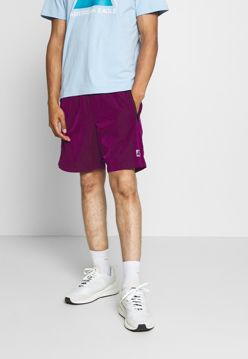 Homme portant un short violet avec des poches latérales, un t-shirt American Eagle bleu clair, des chaussettes blanches et des baskets blanches, debout devant un fond uni.