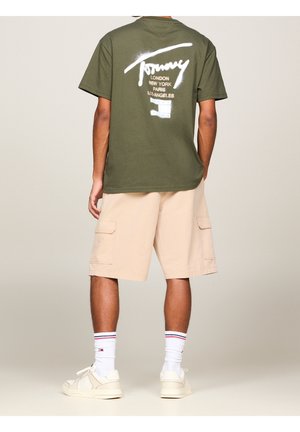 Person iført olivengrøn Tommy grafisk t-shirt, beige cargobukser, hvide sokker med striber og cremefarvede sneakers, stående med ryggen mod seeren.