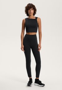 Črn crop top z enim rokavom in detajlom vozla, kombiniran z visokimi črnimi legicami in črnimi športnimi čevlji. Gladka tekstura po celotni površini.