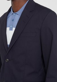 Marinblå blazer med en slät yta, enkel knäppning och bröstficka, över en ljusblå och vit randig pikétröja.