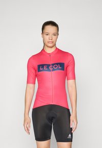 T-skjorte med korte ermer til sykling i lys rosa med en marineblå logo, kombinert med tettsittende svarte shorts med polstret sete. Glatt, elastisk stoff.