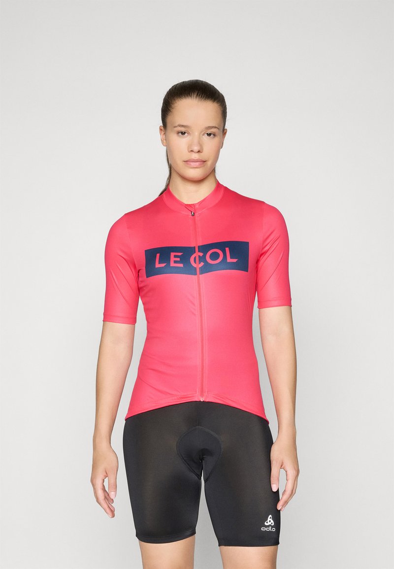 T-skjorte med korte ermer til sykling i lys rosa med en marineblå logo, kombinert med tettsittende svarte shorts med polstret sete. Glatt, elastisk stoff.