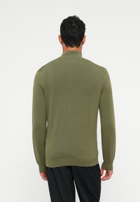 Uomo che indossa un maglione lungo a maniche lunghe verde oliva con collo alto, visto da dietro, abbinato a pantaloni neri.