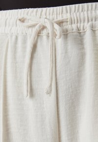Tissu beige clair, texturé, avec une taille élastique et un cordon de serrage. Le tissu semble doux et léger.