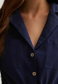 Marinblå linneskjorta med notchkrage, två framfickor och träknappar, med en mjuk textur och skräddarsydd design.