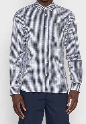 Mann trägt ein eng anliegendes marineblau-weiß kariertes Hemd mit langen Ärmeln und dunkle marineblaue Hose, steht vor einfachem Hintergrund.