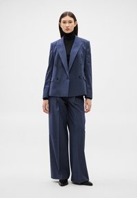 Sieviete tumszilu dubultpogas žaketi un platām biksēm pār melnu augstu apkakli, stāvot pret vienveidīgu gaišu fona.