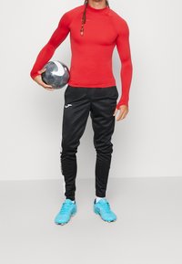 Czerwona, długorękawowa sportowa koszulka, czarne spodnie dresowe z białymi paskami oraz niebieskie korki piłkarskie. Trzyma czarno-białą piłkę nożną.