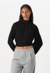 Top nero plissettato cropped con collo alto e maniche lunghe e larghe, abbinato a pantaloni sartoriali grigio chiaro a righe.