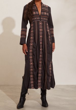 Mujer vestida con un vestido largo negro con bordados geométricos intrincados en vertical y horizontal, combinado con botas negras de tacón.