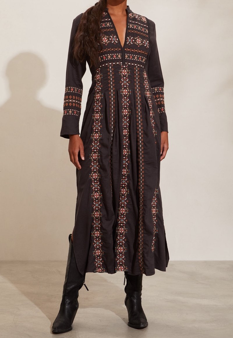 Femme portant une longue robe noire ornée de broderies géométriques verticales et horizontales complexes, associée à des bottes noires à talons.