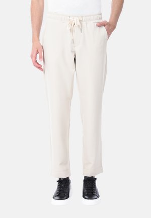 Uomo che indossa pantaloni avorio con coulisse e sneakers nere, in piedi con una mano in tasca su uno sfondo semplice.