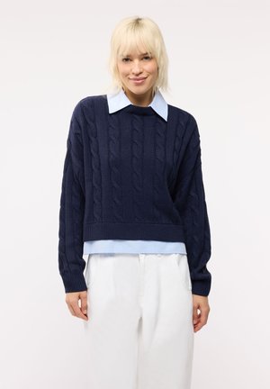 Donna con capelli biondi che indossa un maglione blu navy lavorato a trecce sopra una camicia celeste con colletto e pantaloni bianchi su uno sfondo semplice.