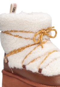 Stivale marrone e crema con una parte superiore soffice, caratterizzato da un dettaglio di laccio giallo e una base liscia simile alla pelle. Tra gli accessori è presente un bottone decorativo.