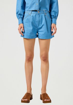 Shorts de mezclilla azul con cintura alta, que cuentan con bolsillos delanteros y un dobladillo sin terminar. Combinados con sandalias de tacón de ante marrón.