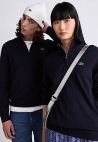 Dva pojedinca nose tamnoplave dukserice sa patentnim zatvaračem i rebrastim manžetama. Dukserice imaju mali zeleni logo. Jedna osoba nosi belu kapu i srebrnu torbu.