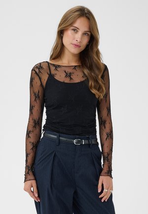 Jeune femme aux longs cheveux portant un haut noir transparent en dentelle florale à manches longues et un pantalon taille haute bleu marine à fines rayures avec une ceinture noire.