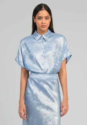 Femme aux longs cheveux foncés portant un chemisier court à manches courtes bleu clair brillant et une jupe taille haute assortie, sur fond gris.