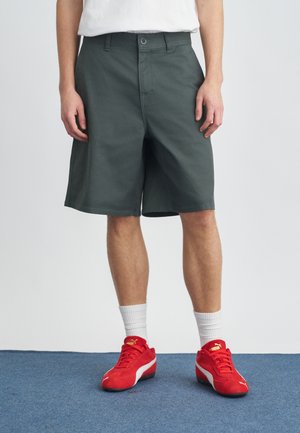 Tmavozelené šortky z bavlnenej zmesi s voľným strihom, bočnými vreckami a zapínaním na zips, nosené s červenými športovými topánkami.