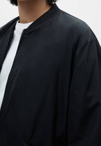 Chaqueta bomber negra con cuello acanalado, cierre de cremallera y textura suave, llevada sobre una camisa blanca. Detalles incluyen bolsillos laterales.