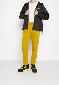 Černá lehká bunda s kapucí, bílé tričko, žluté tepláky a černé tenisky se žlutými detaily, stojící na bílém pozadí.