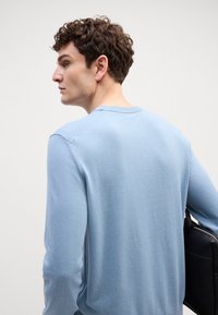 Ανοιχτοί μπλε πλεκτός πουλόβερ τύπου crewneck με λεία υφή, ριχτή λαιμόκοψη και ράγλες μανίκια. Το μοντέλο κρατάει μια μαύρη τσάντα στον ώμο.
