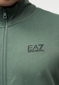 Πράσινο μπουφάν με φερμουάρ κατασκευασμένο από λείο ύφασμα, με ψηλό γιακά, μαύρο λογότυπο EA7 Emporio Armani στο στήθος και φερμουάρ σε αντίθετο χρώμα.