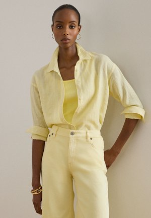 Γυναίκα που φοράει ανοιχτό κίτρινο πουκάμισο με κουμπιά πάνω από κίτρινο τοπ χωρίς μανίκια, συνδυασμένο με ψηλόμεσο κίτρινο παντελόνι, χρυσούς κρίκους σκουλαρίκια και βραχιόλια.