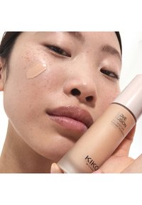 Gros plan sur une personne avec du fond de teint sur la joue tenant un flacon de fond de teint KIKO MILANO Love Fusion 24H Moisture Radiant.