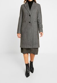 Abrigo gris de espiga con solapa de muesca, cierre de un solo botón y dos bolsillos laterales, llevado sobre un vestido oliva hasta la rodilla y botas negras puntiagudas.