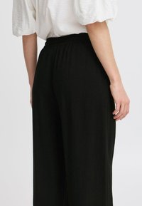 Schwarze weit geschnittene Hose mit elastischem Bund, weicher Textur und glatter Oberfläche, kombiniert mit einem weißen Oberteil mit Puffärmeln.