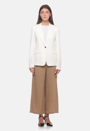 Donna in piedi, rivolta in avanti, indossa un blazer bianco, una maglietta bianca, pantaloni beige a gamba larga e tacchi neri su uno sfondo bianco semplice.