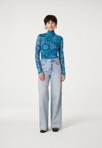 Langarm, hoge hals top in helderblauw met bloemen- en zonafdrukken. In combinatie met lichtblauwe wijde jeans en zwarte schoenen.