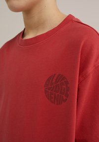 Sarkans kokvilnas T-krekls ar apaļu kakla izgriezumu un īsām piedurknēm. Uz kreisā krūšu daļas ir melns apļa grafiskais logo ar viendabīgu tekstūru.