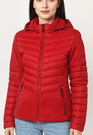 Mujer con una chaqueta acolchada roja con capucha, que tiene cremallera frontal y bolsillos con cremallera, combinada con unos vaqueros de color azul claro.