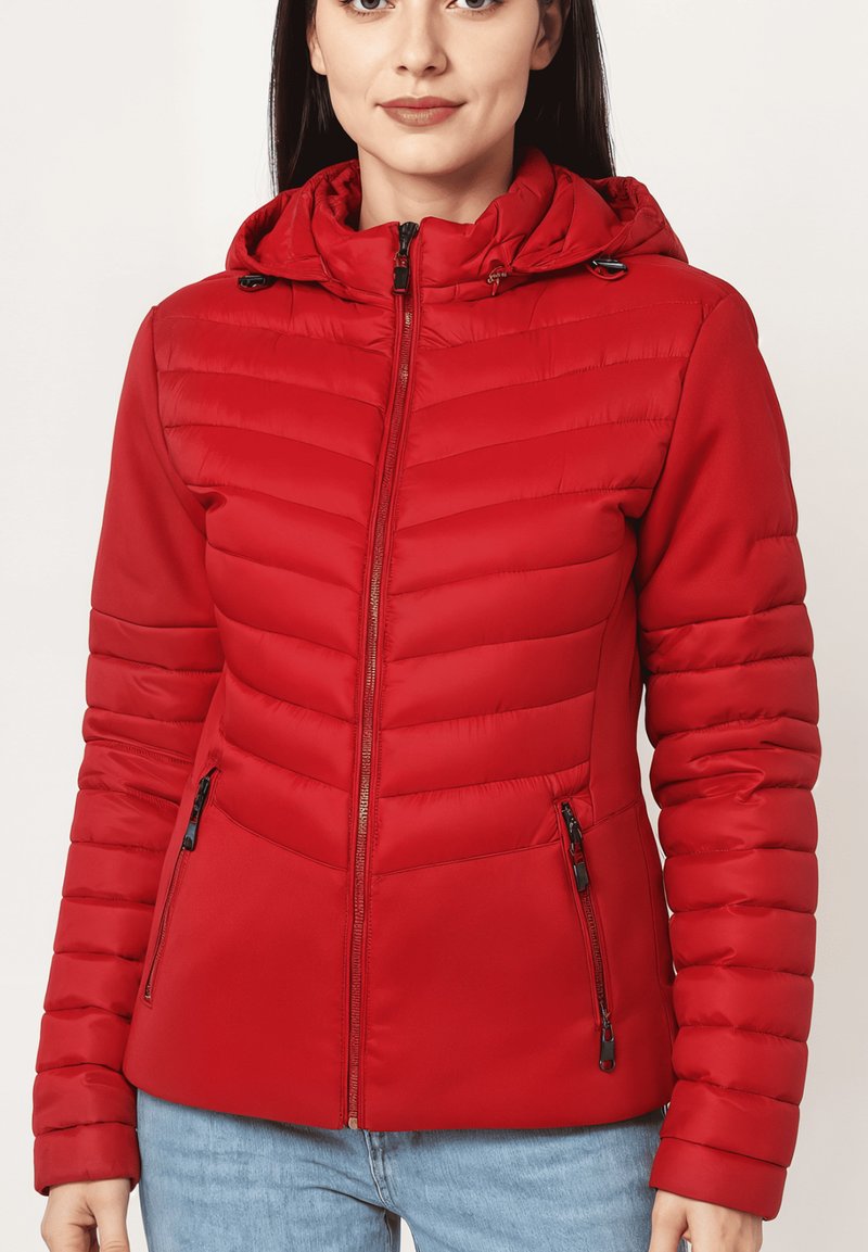 Mujer con una chaqueta acolchada roja con capucha, que tiene cremallera frontal y bolsillos con cremallera, combinada con unos vaqueros de color azul claro.