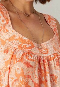 Top de color naranja con patrón de paisley, escote en forma de uno, cuerpo fruncido y textura en capas. Presenta un collar de oro sencillo con un colgante ovalado.