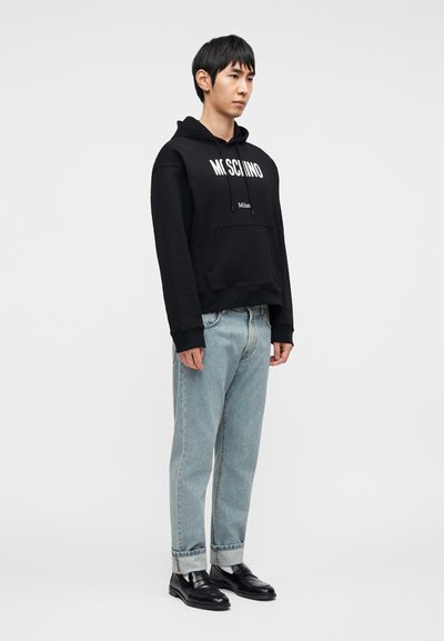 Μαύρο φούτερ με κουκούλα με λευκή επιγραφή "MOSCHINO" και "Milan", εμπρόσθια τσέπη, σε συνδυασμό με ανοιχτόχρωμο μπλε τζιν και μαύρα δερμάτινα παπούτσια. Μινιμαλιστικός σχεδιασμός.