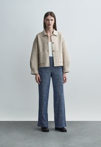 Nő áll, bézs gombos dzsekit visel nagy zsebekkel, fehér felsőt, texturált kék bő szárú nadrágot és fekete cipőt.