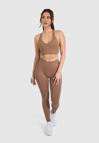 Un reggiseno sportivo a coste marrone e leggings a vita alta, entrambi con una texture liscia. Indossati con scarpe da ginnastica bianche, su uno sfondo bianco.