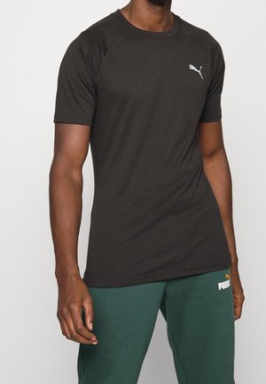 Homme portant un t-shirt noir ajusté à manches courtes Puma et un pantalon de jogging vert Puma, debout avec un bras détendu le long du corps.