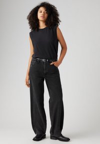 Zwarte mouwloze top, hoge taille zwarte wijde jeans, zwarte riem en enkelboots. Glad materiaal, comfortabele pasvorm, minimale details.