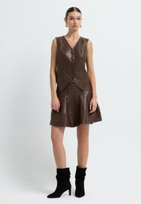 Gilet sans manches en faux cuir marron avec cinq boutons, associé à une jupe évasée assortie. Le modèle porte des bottes noires à talons plats.