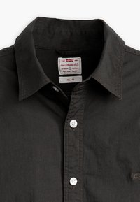 Camisa negra ajustada hecha de una tela suave. Presenta un cuello estándar, botones blancos y una etiqueta de Levi's en el escote.