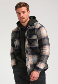 Gequilted geruite shirtjack in beige, marineblauw en grijze tinten; voorzien van twee borstzakken en zwarte knoopsluitingen; gedragen over een groene hoodie.