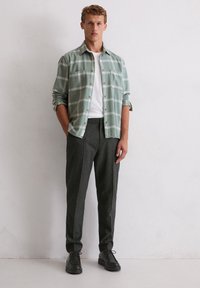 Jonge man in grijs geblokte broek, wit T-shirt en groen geblokt overhemd met opgerolde mouwen tegen een witte muur.