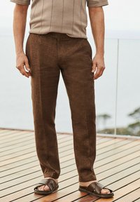 Mann in braunen Leinenhosen, braunen Sandalen und einem kurzärmligen taupefarbenen Hemd steht auf einer Holzterrasse mit Glasgeländer.