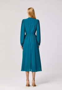 Robe teal à manches longues avec une taille cintrée et une jupe fluide. Comprend des plis subtils et une fermeture éclair cachée dans le dos.
